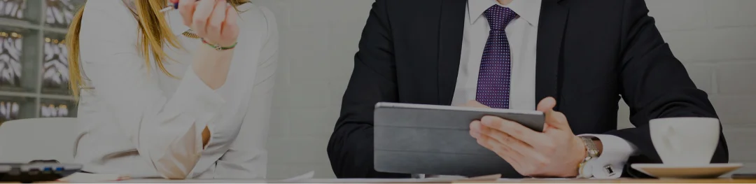 A man in a suit and tie is seated at a desk, focused on a tablet in front of him.