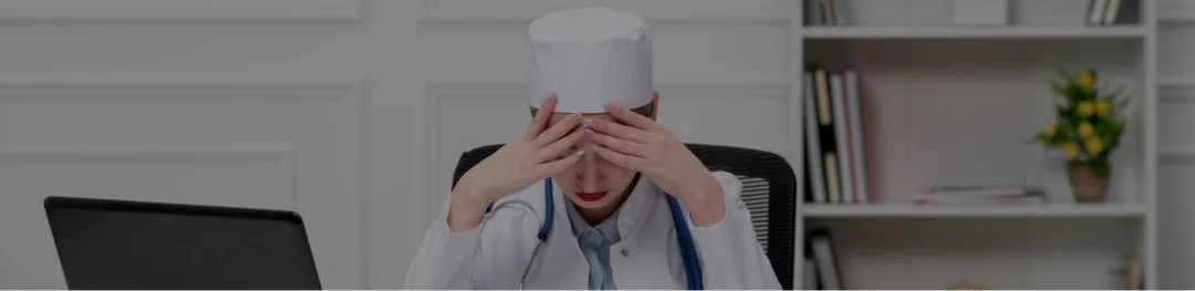 A woman in a white coat conceals her face with her hands, expressing a moment of contemplation or distress.