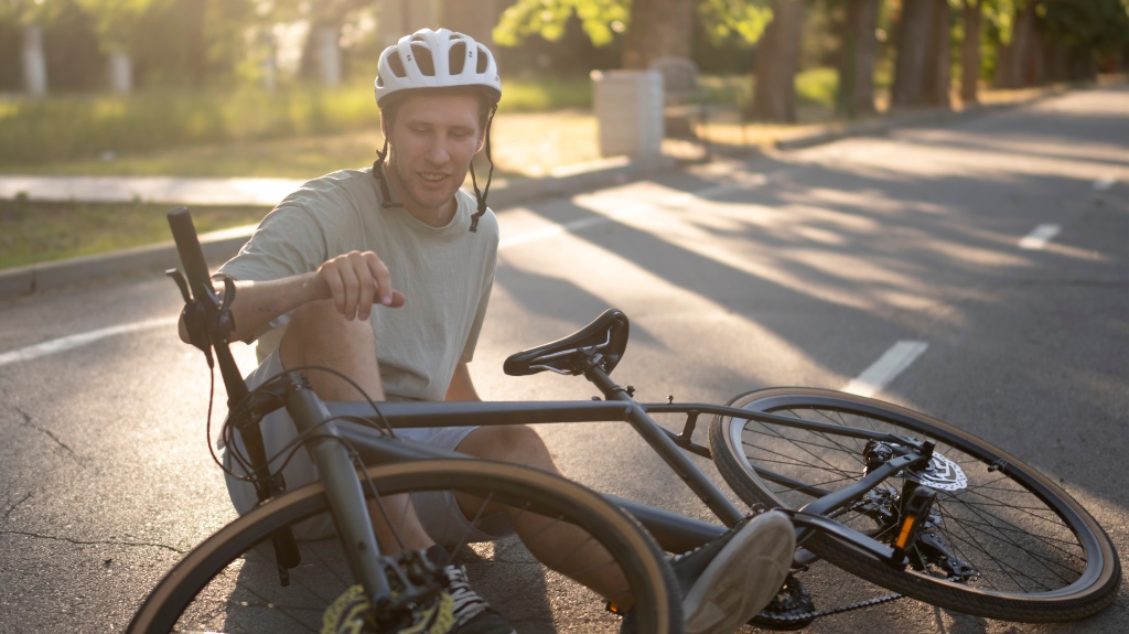 man wearing a helmet sits on the asphalt of a city street, his leg injured, after a bicycle crash